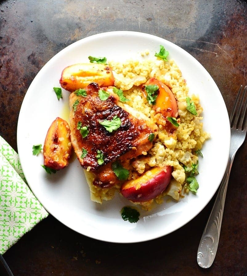 Top down view of chicken with peach wedges on a bed of bulgur on top of white plate, with fork to the right and green cloth to the left.