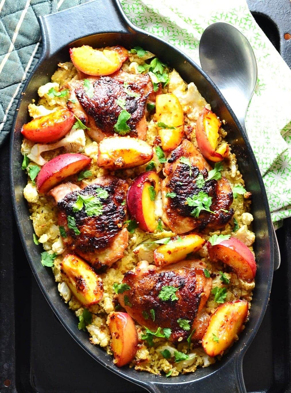 Top down view of crispy chicken with peaches on a bed of cauliflower and bulgur inside black oval casserole dish, with spoon on top of green cloth to the right.