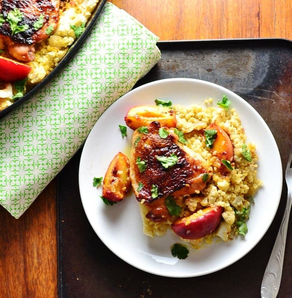 Top down view of honey chicken and peach wedges with bulgur on top of white plate, with fork to the right. green cloth to the left and partial view of chicken casserole in top left corner.