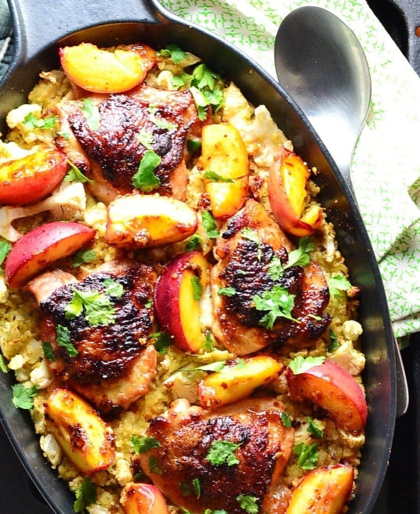 Top down partial view of crispy chicken with peaches on a bed of cauliflower and bulgur inside black oval casserole dish, with spoon on top of green cloth to the right.