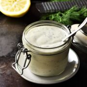 Healthy ranch dressing in open jar with spoon, lemon and herbs in background.