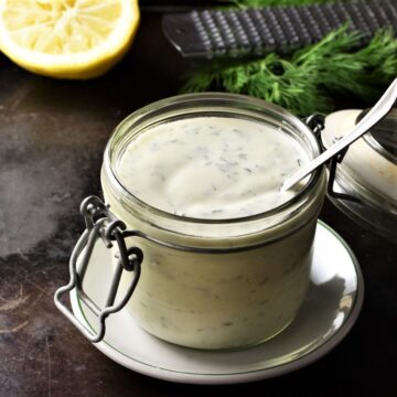 Healthy ranch dressing in open jar with spoon, lemon and herbs in background.
