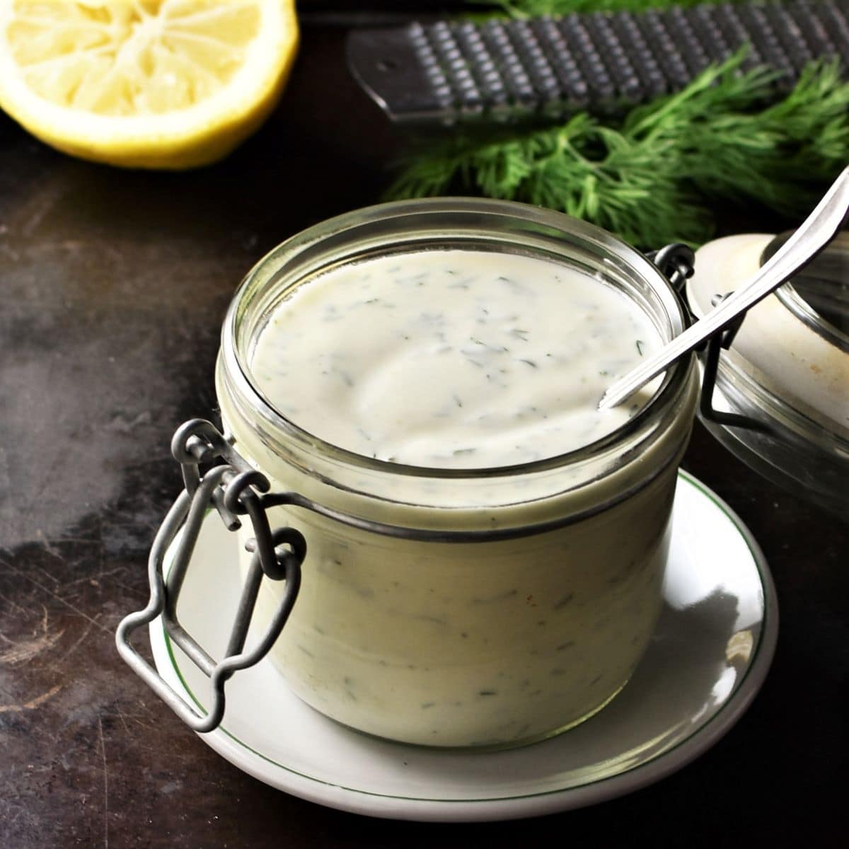 Healthy ranch dressing in open jar with spoon, lemon and herbs in background.