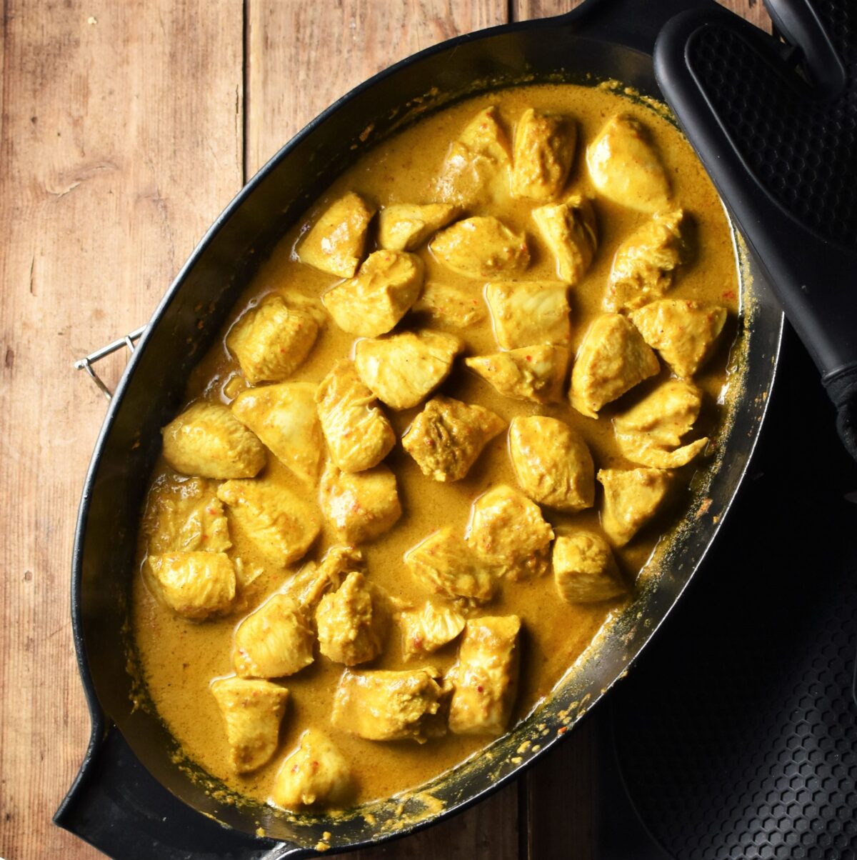 Chicken pieces in curry sauce in black oval skillet.