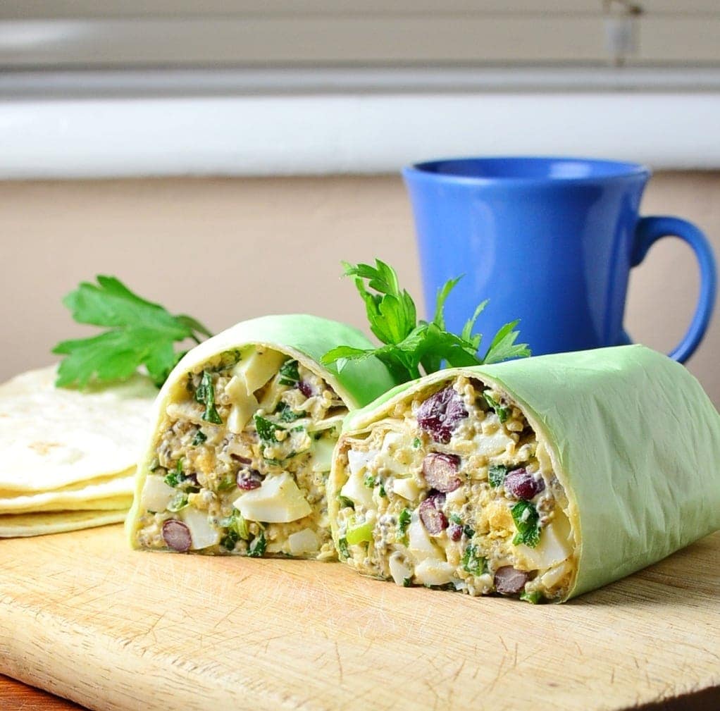 Side view of halved egg salad wrap on top of wooden board with parsley leaves and blue cup in background.