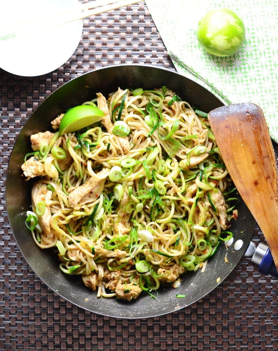 Hoisin chicken stir fry in black wok with wooden spatula, bowl with chopsticks and lime on green cloth.