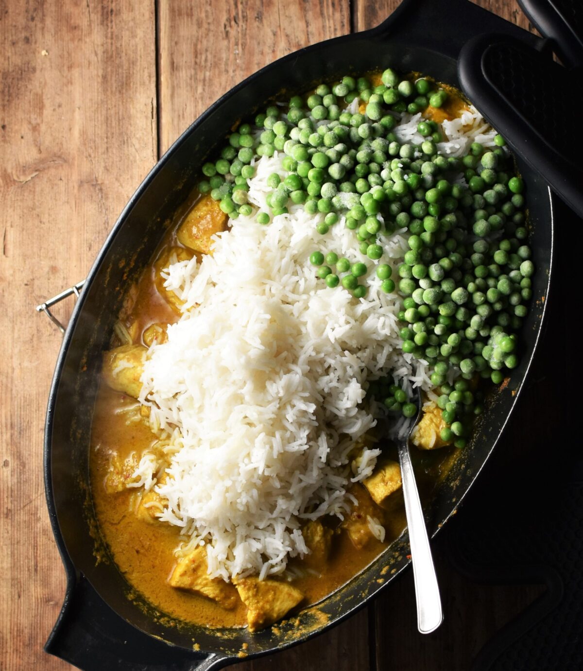 Cooked rice and peas on top of creamy chicken curry with spoon in large black oval pan.