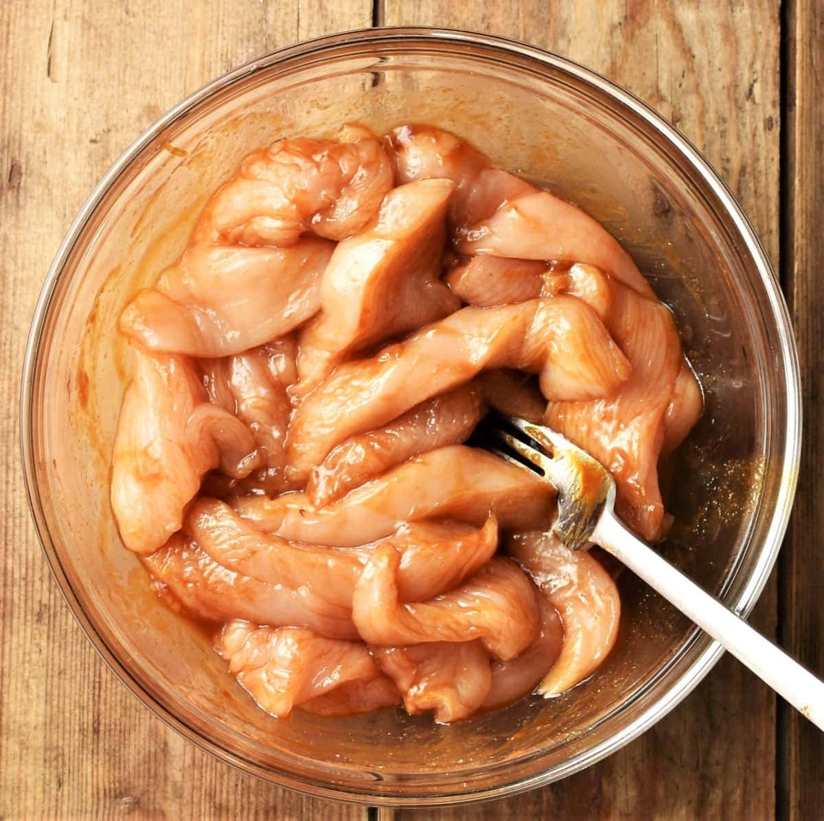 Chicken strips in hoisin marinade in mixing bowl with fork.