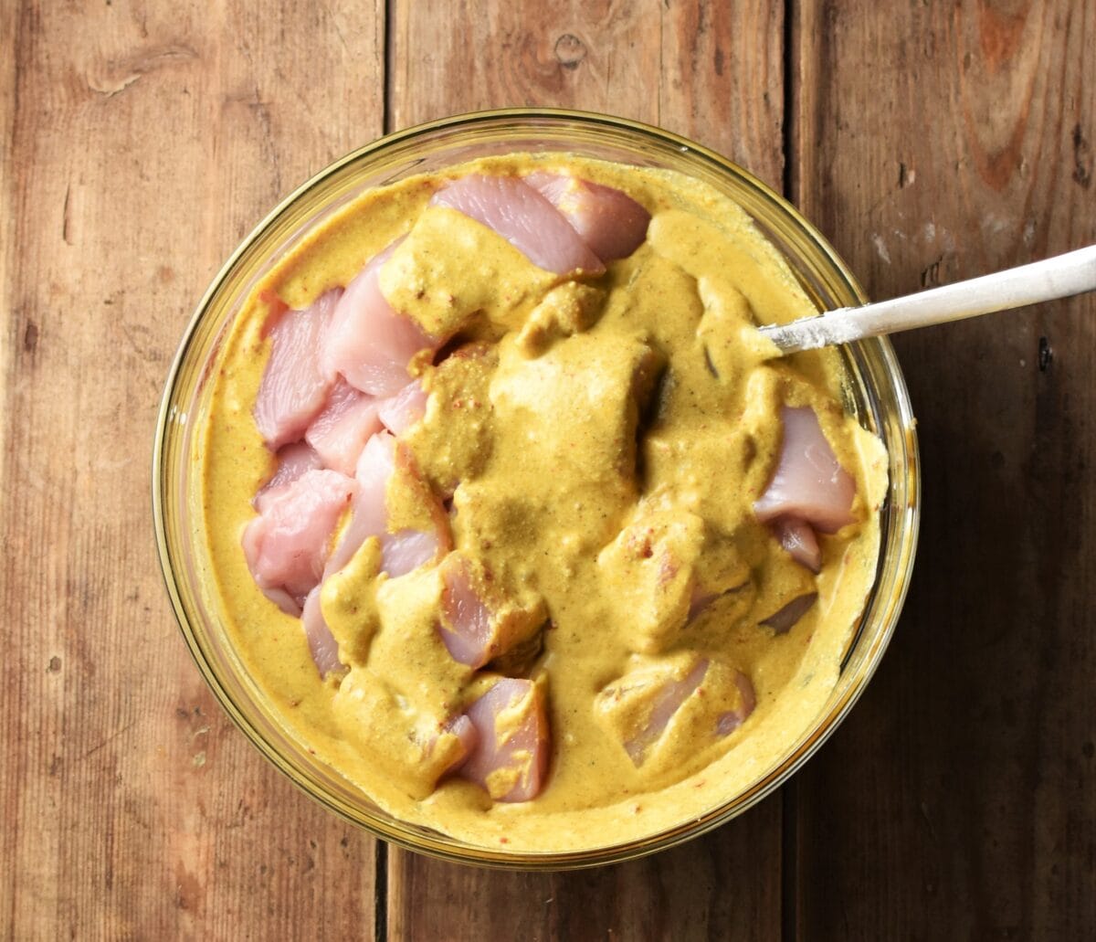 Raw chicken pieces in curried creamy marinade in mixing bowl with spoon.