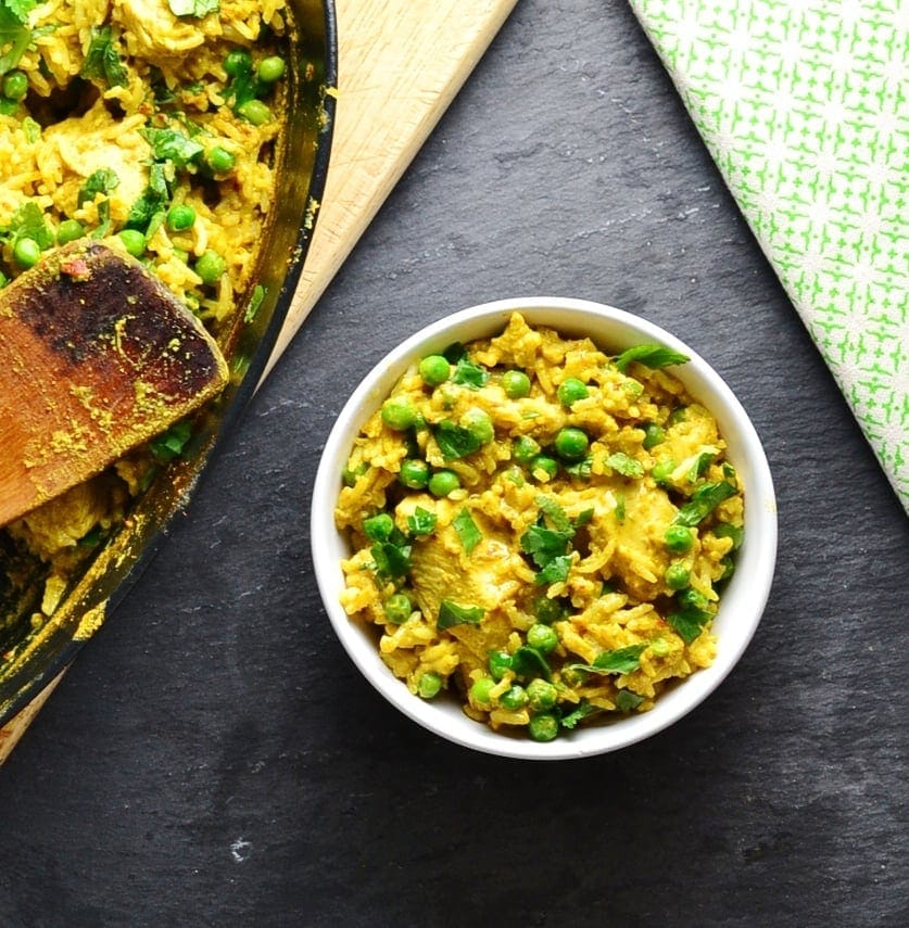 Top down view of curried chicken rice casserole in black oval dish on top of wooden board, with wooden spoon in bottom right corner.