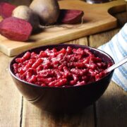 Beetroot pasta in purple bowl with spoon, cloth and raw beets in background.