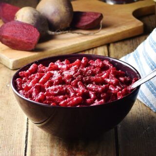 Beetroot pasta in purple bowl with spoon, cloth and raw beets in background.