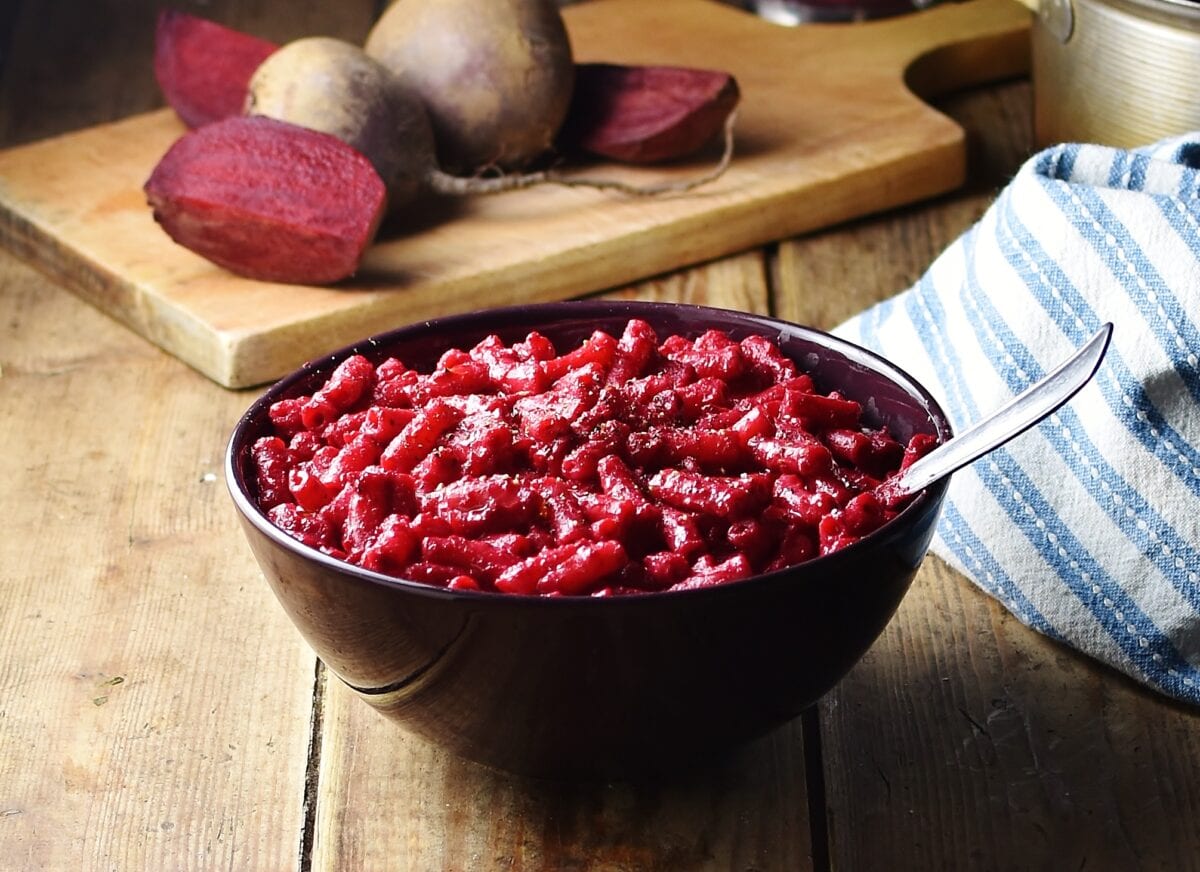 Beetroot pasta in purple bowl with spoon, blue-and-white stripy cloth and raw beetroot on wooden board in background.