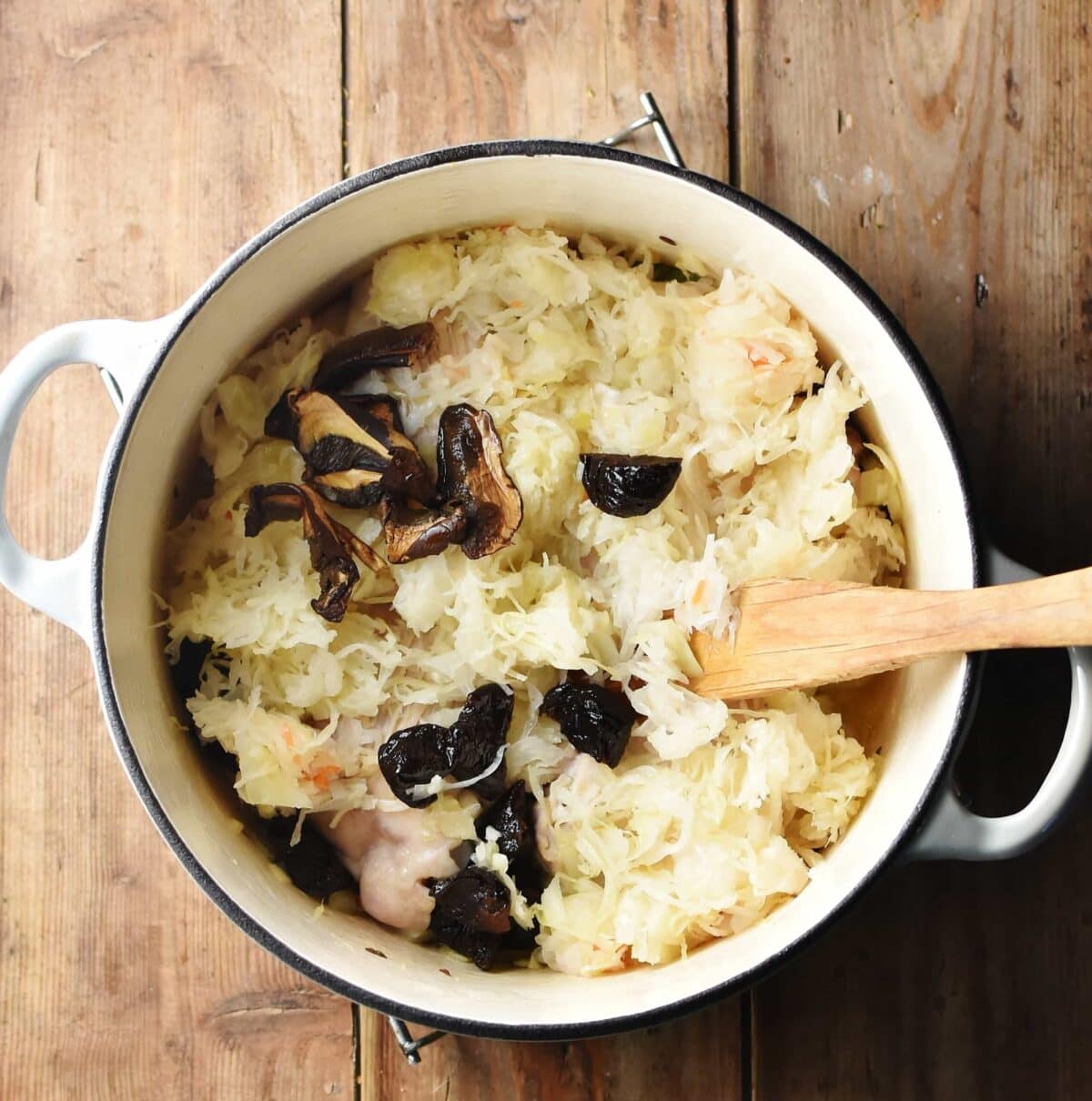 Sauerkraut, prunes and dried mushrooms in large white pot with wooden spoon.