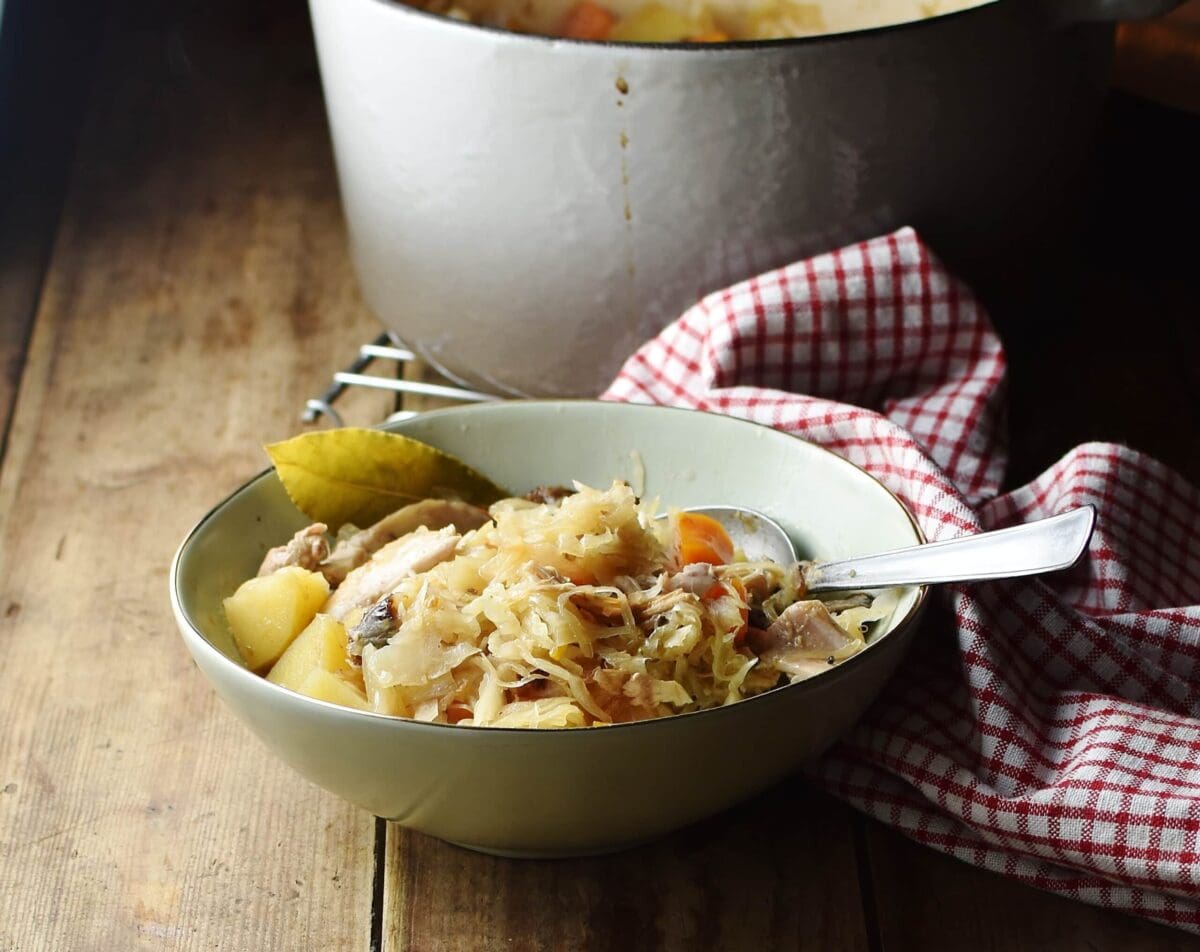 Side view of sauerkraut, chicken and potato casserole in green bowl with spoon, red-and-white cloth and large white pot in background.