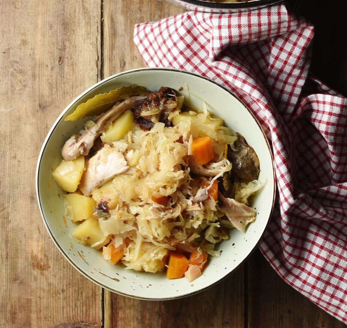 Sauerkraut, chicken, potato, carrot stew in green bowl with red-and-white checkered cloth to the right.