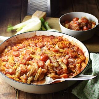 Fussili pasta with sliced fennel and tomato sauce in large white casserole dish with spoon, pasta and sauce in bowl, halved fennel and grater in background.