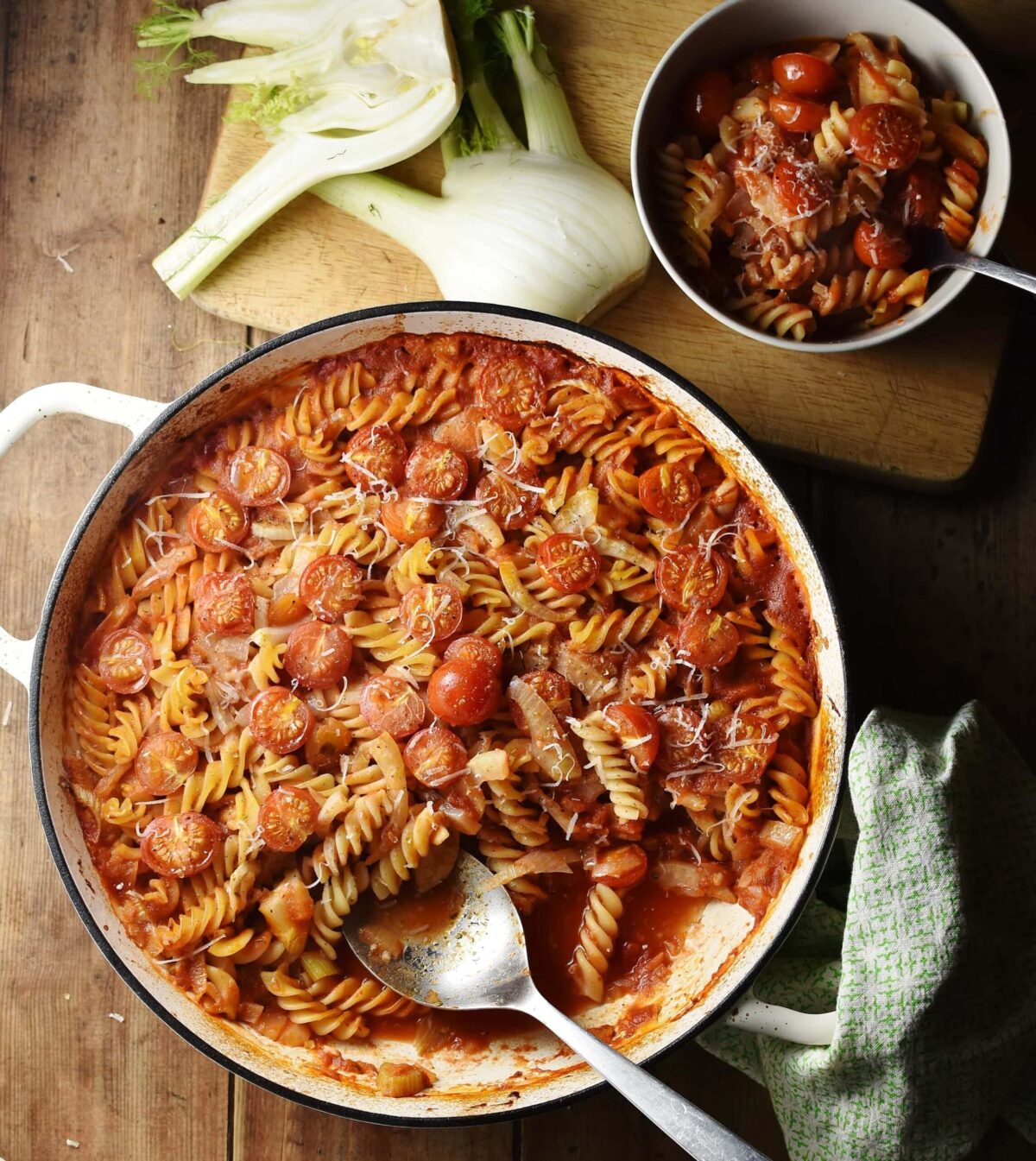 Top down view of pasta with tomatoes and fennel in large shallow white dish with spoon, raw fennel and pasta bake in small bowl in background.