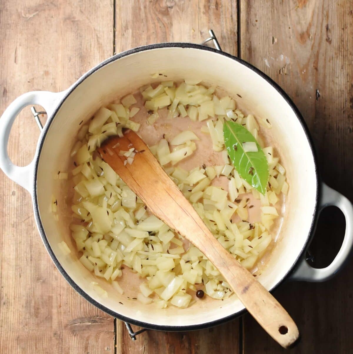 Chopped onion and spices in large white pot with wooden spatula.
