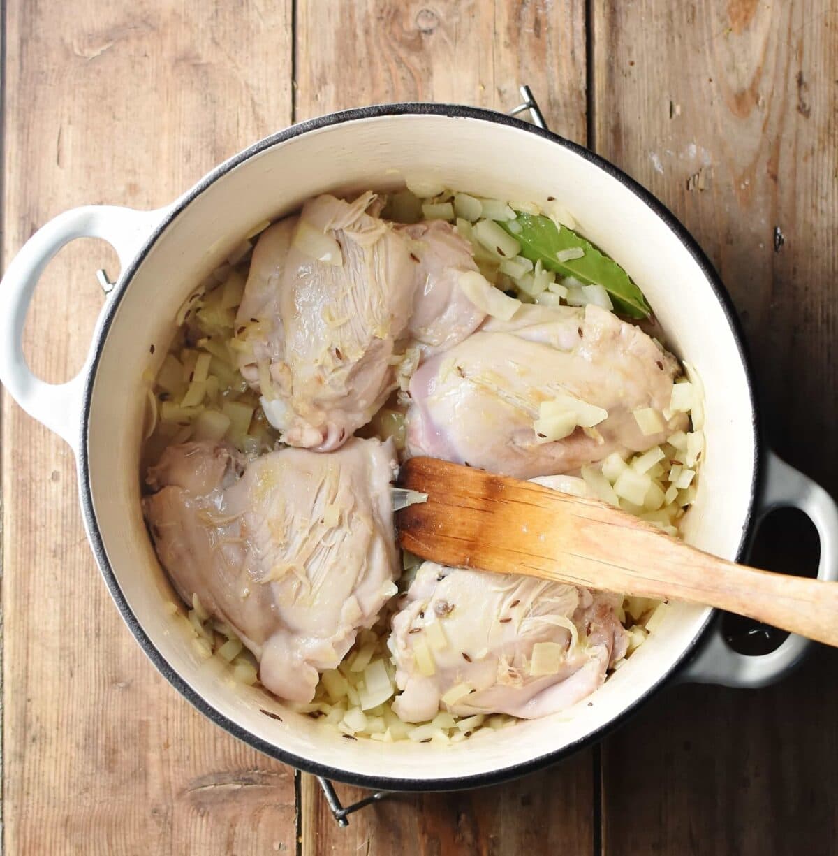 4 chicken thighs with chopped onions in large white pot with wooden spatula.