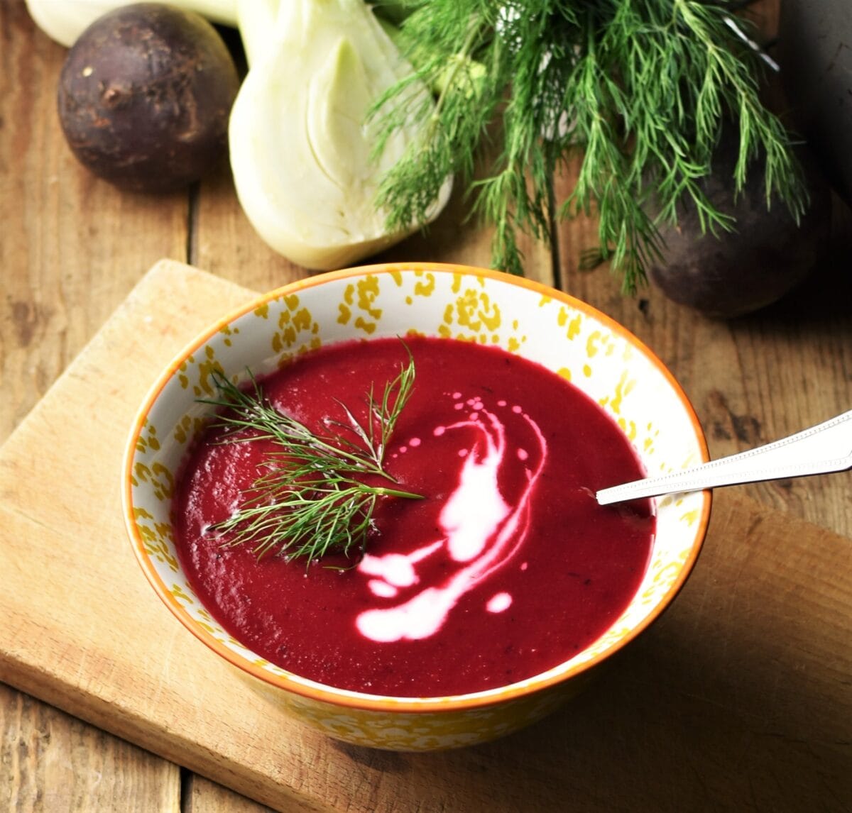 Creamy beetroot soup with dill and yogurt in yellow patterned bowl with spoon on chopping board with beet, fennel and dill in background.