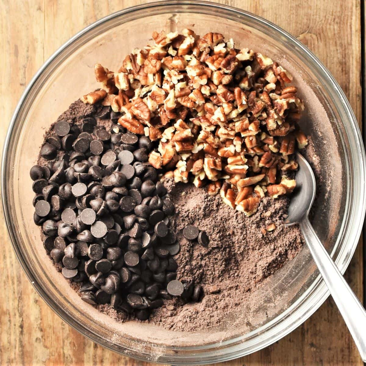 Cocoa mixture with chocolate chips and chopped pecans in mixing bowl with spoon.