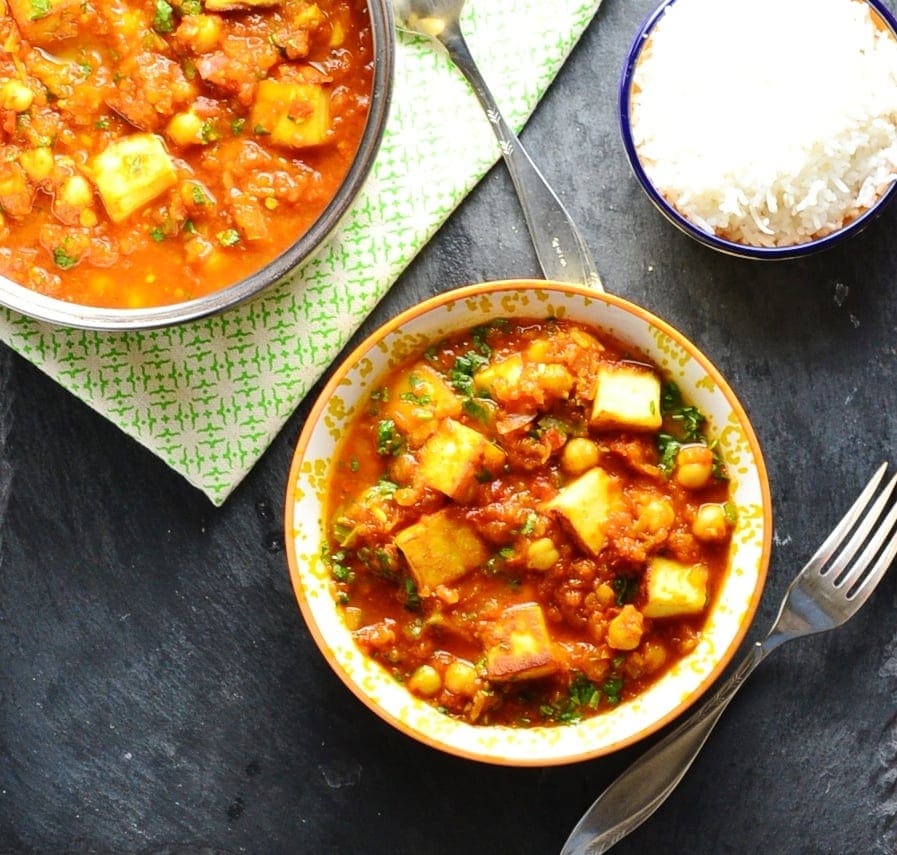 Top down view of vegetarian curry with paneer in white-and yellow bowl and saucepan, with fork, green cloth and bowl of rice.