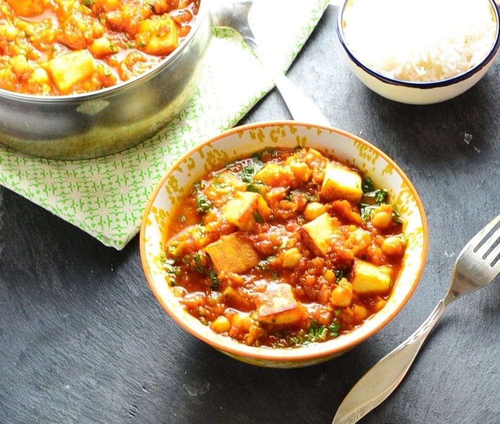 Vegetable curry in white-and-yellow bowl with saucepan and bowl of rice in background and fork to right.