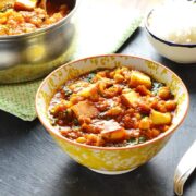 Vegetable curry with paneerin white-and-yellow bowl with saucepan and bowl of rice in background and fork to right.