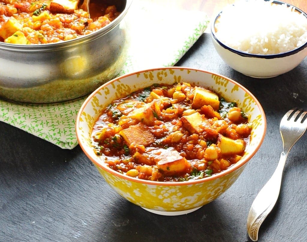 Vegetarian curry with paneer in white-and-yellow bowl with saucepan and bowl of rice in background and fork to right.