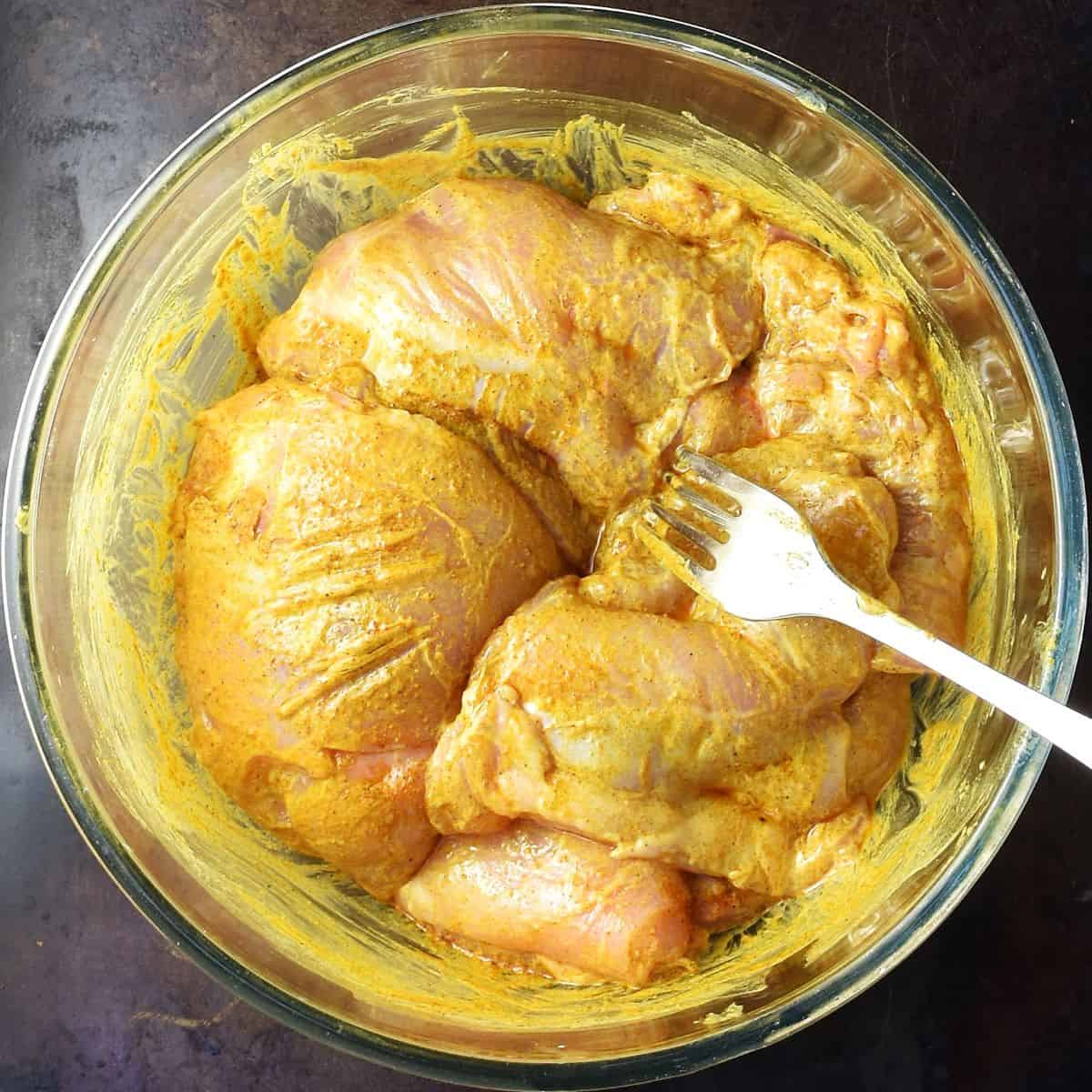 Skinless chicken thighs coated in spices with fork in glass bowl.