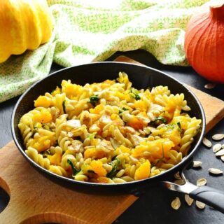 Side view of chopped pumpkin with fussili pasta, arugula, garlic slices and nuts on top of wooden board, with green cloth and pumpkin in background.