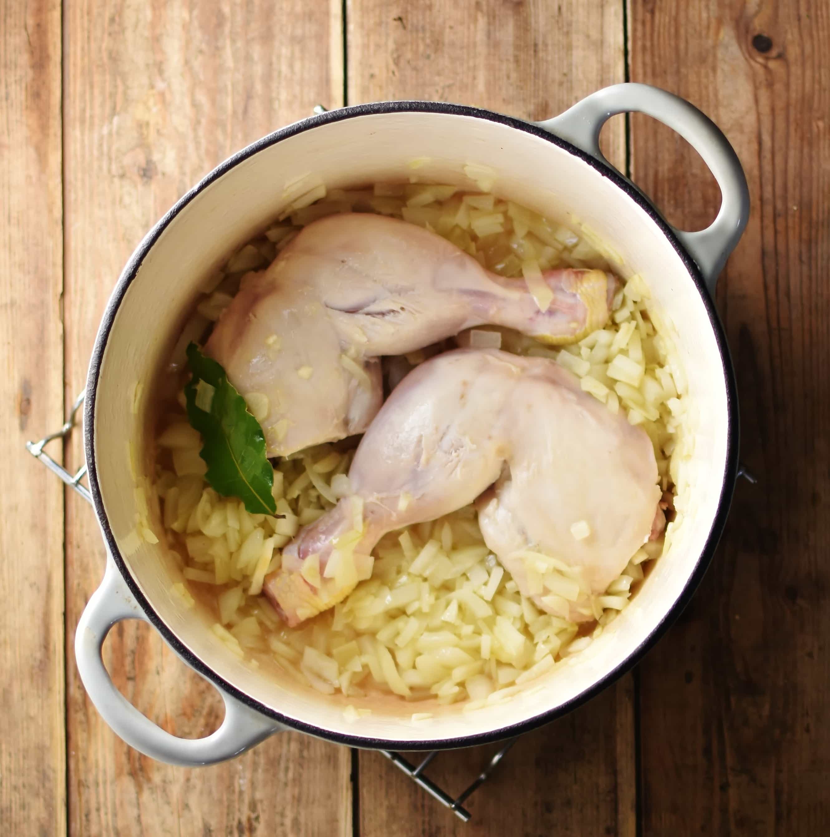 2 chicken leg quarters with bay leaf and onions in white pot.