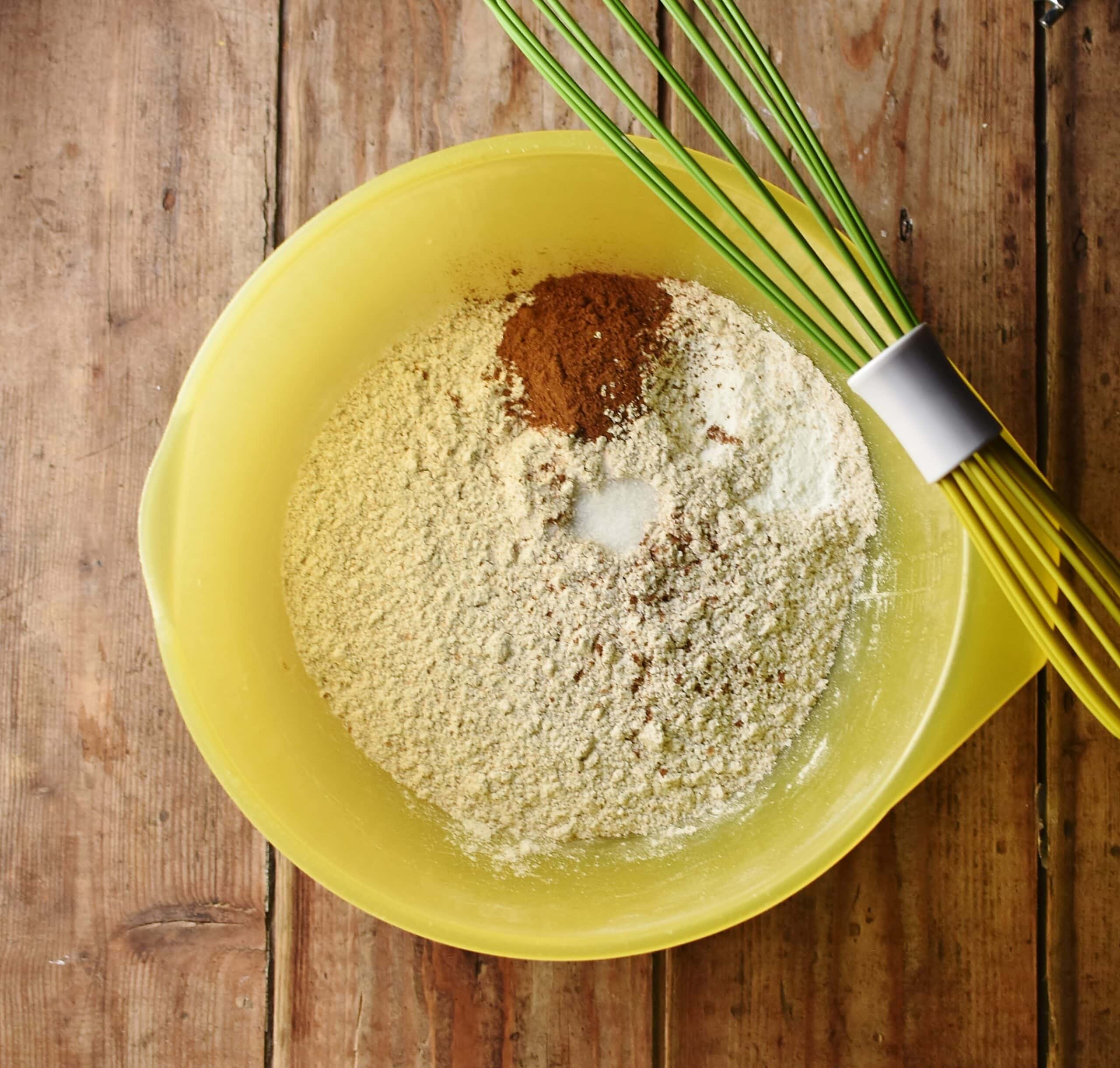 Flour and spice mixture in large yellow bowl with green whisk.