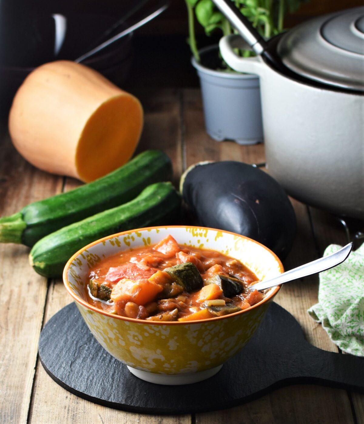 Side view of vegetable stew in yellow patterned bowl with spoon, zucchini, eggplant, squash and pot in background.