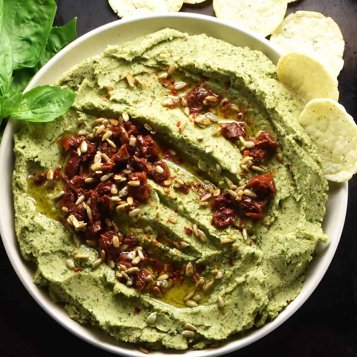 Top down view of creamy broccoli dip with sundried tomatoes and seeds on white plate with herbs and crisps in background.