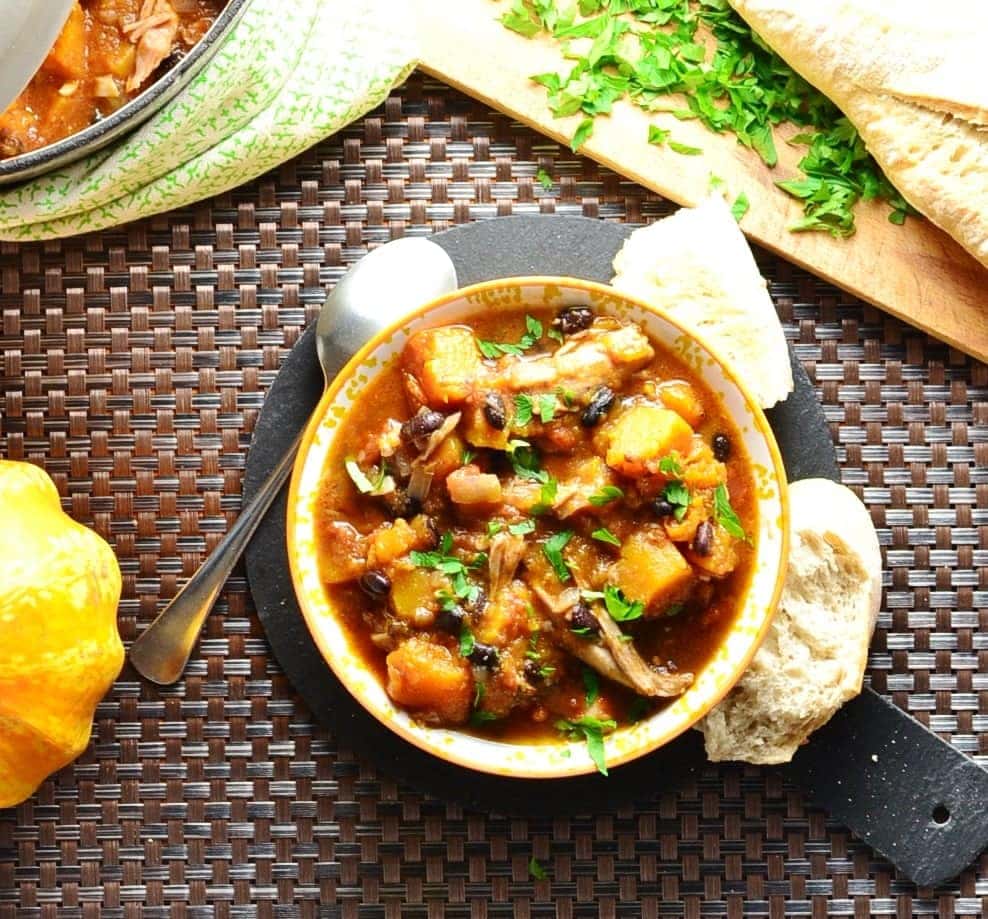 Chicken butternut squash casserole in yellow-and-white bowl with spoon, roll, and partial view of casserole dish, wooden board with herbs and squash.