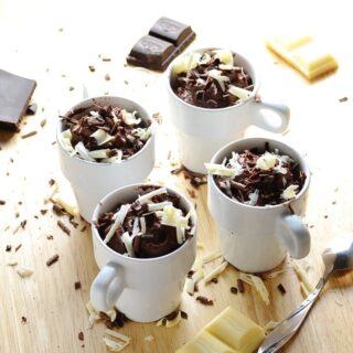 Cheesecake pots with white and dark chocolate shavings, chocolate pieces and spoon on wooden surface.