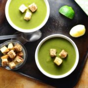 Top down view of split pea soup in 2 white bowls with croutons, lime wedges, spoons and dish of croutons on oven tray.