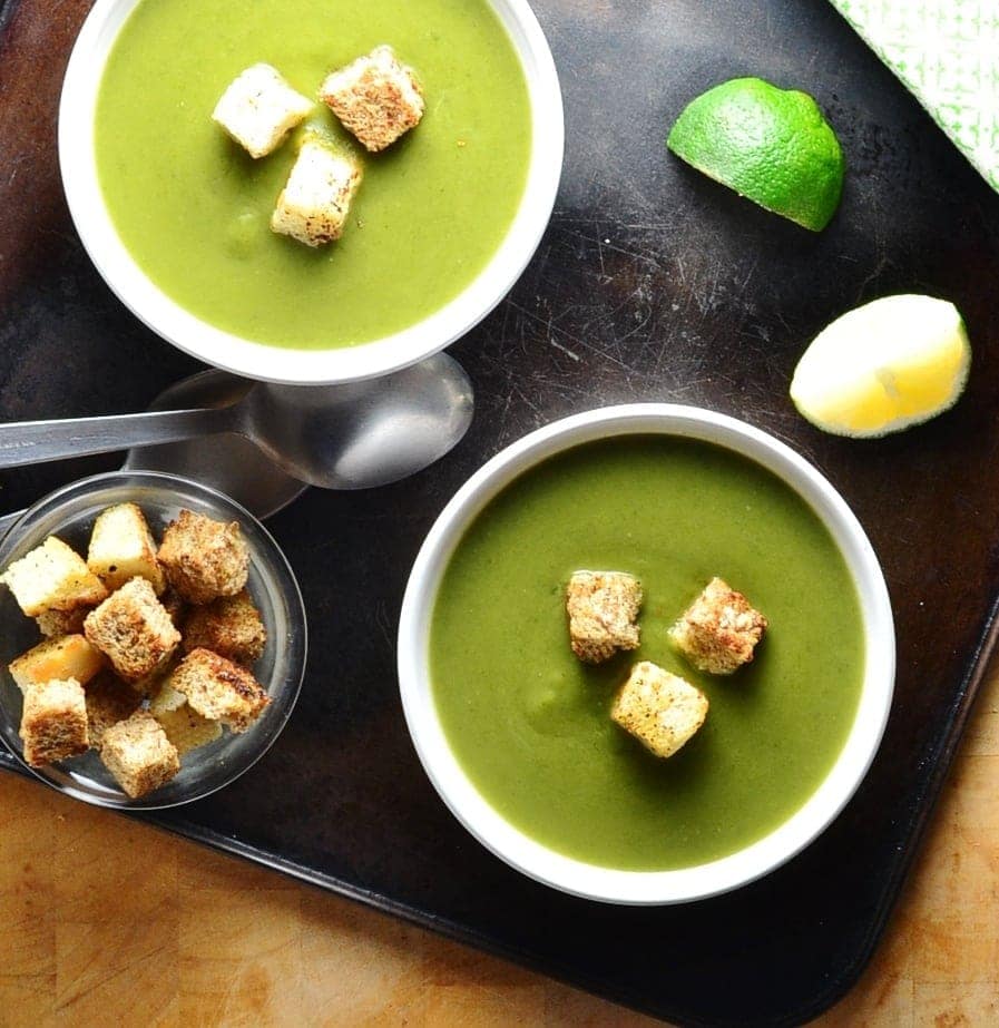 Top down view of green pea soup in 2 white bowls with croutons, lime wedges, spoons and dish of croutons on oven tray.