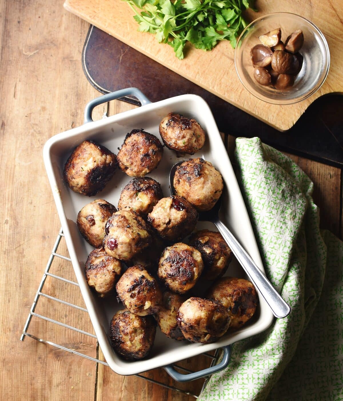 Top down view of turkey stuffing balls in rectangular dish with spoon, with green cloth to the right and chestnuts in small dish and herbs on top of wooden board at the top.
