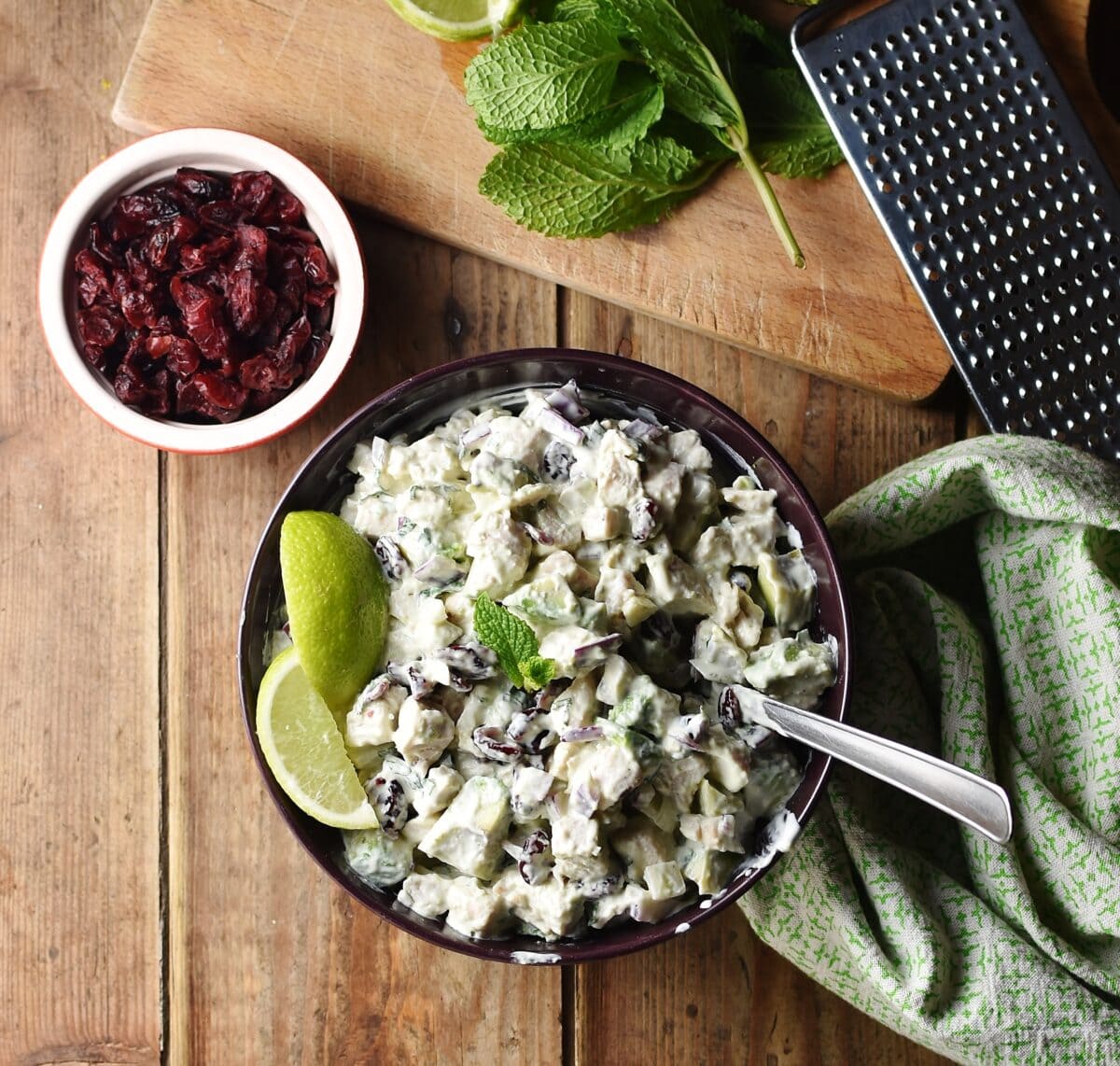 Top down view of creamy chopped turkey, avocado and dried cranberries with lime wedges in purple bowl with spoon and green cloth to the right, dried cranberries in white dish, mint leaves and fruit zester in the background.