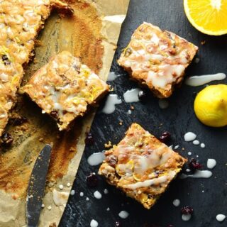Top down view of fruit cake slices on top of grey surface with halved lemon and orange, as well as knife.