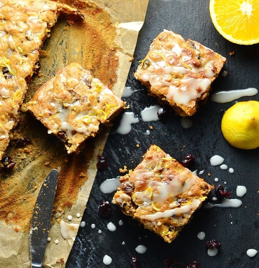 Fruit cake squares on top of dark table and parchment paper, with orange, lemon and knife.