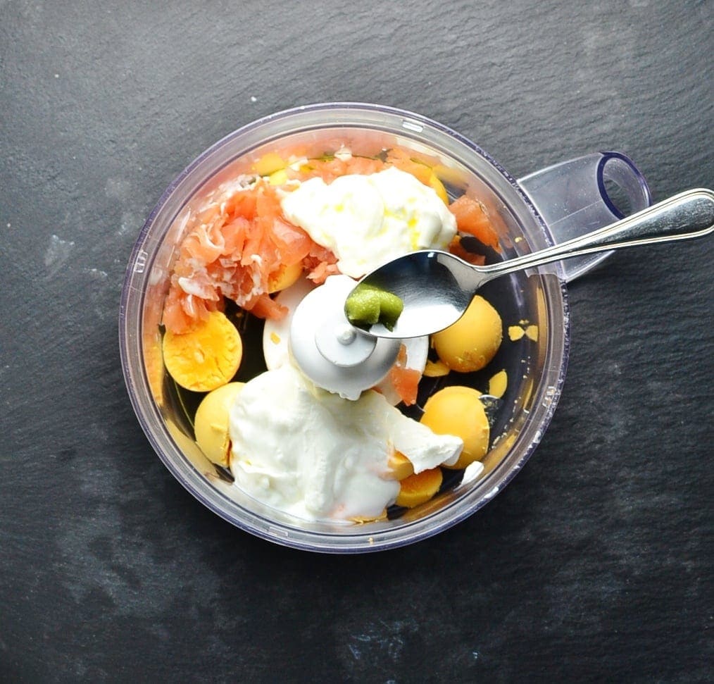 Smoked salmon deviled eggs ingredients in blender bowl with spoon on grey surface.