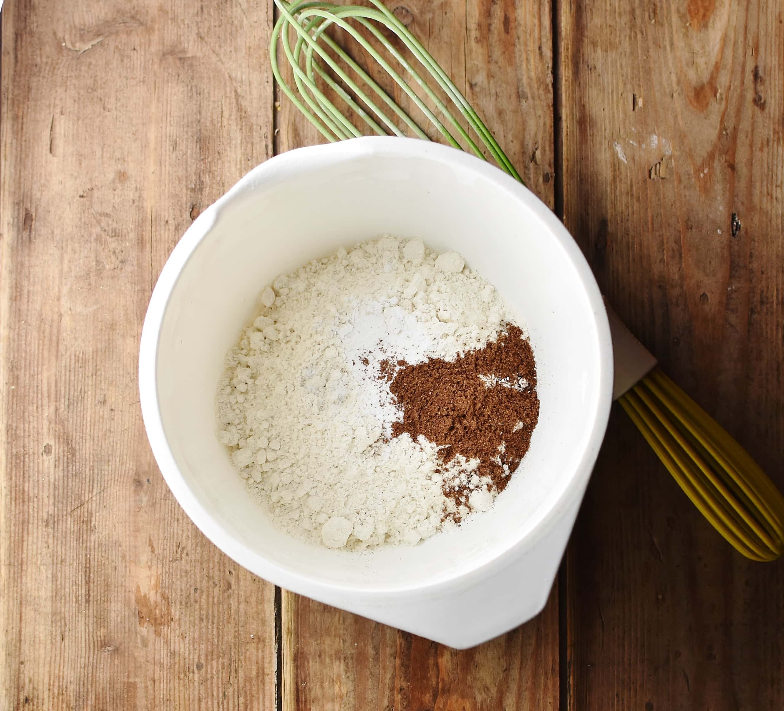 Flour and spices in white bowl with green whisk to the right.