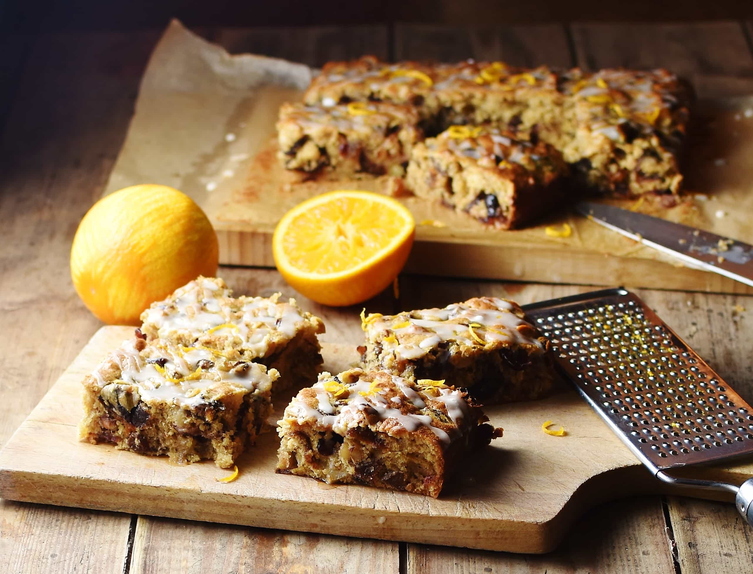 Slices of fruit cake on top of wooden board with zester to the right and oranges and more fruit cake in background.