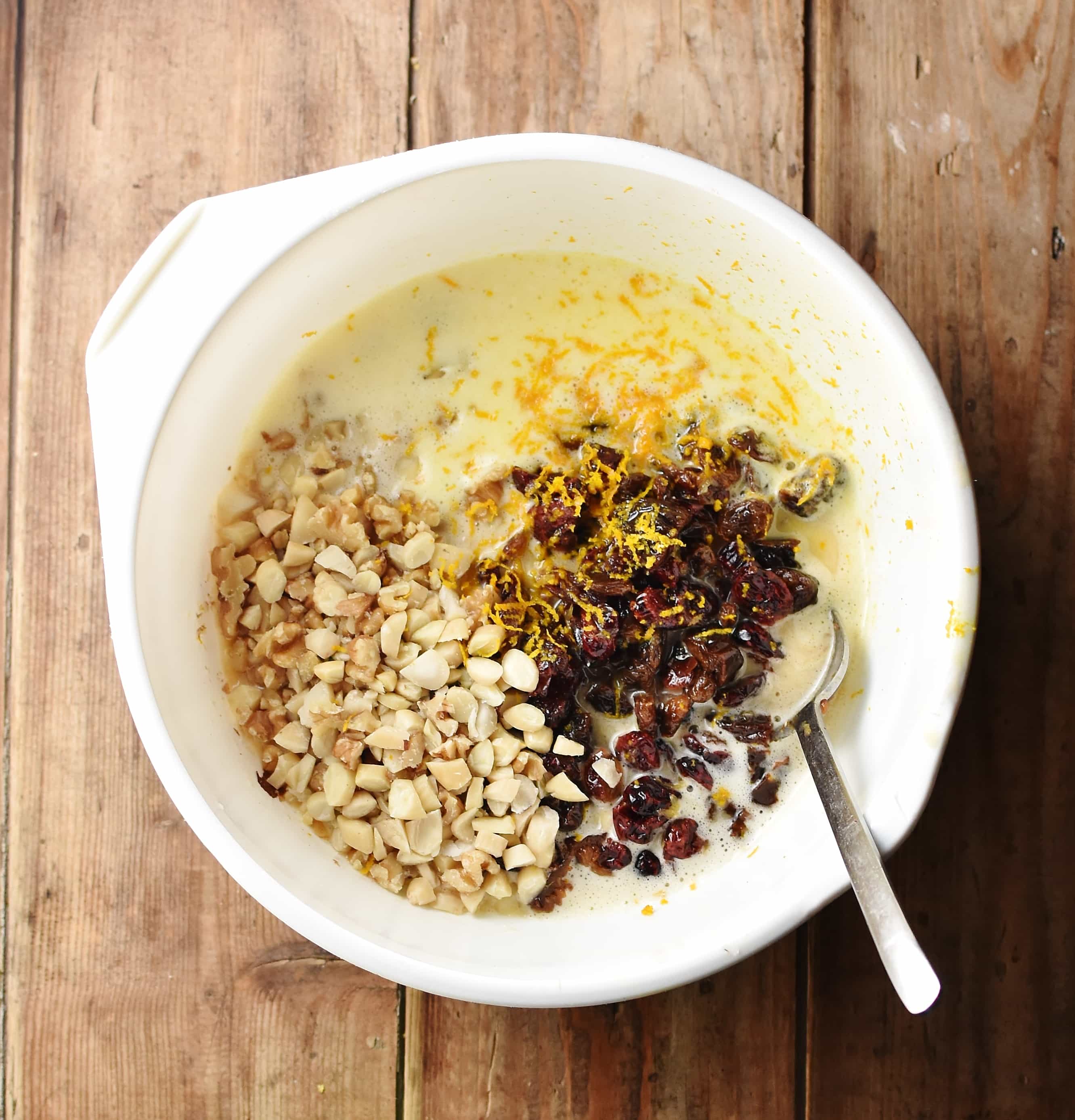 Top down view of nuts, dried fruit, egg mixture and orange zest in large white bowl with spoon.