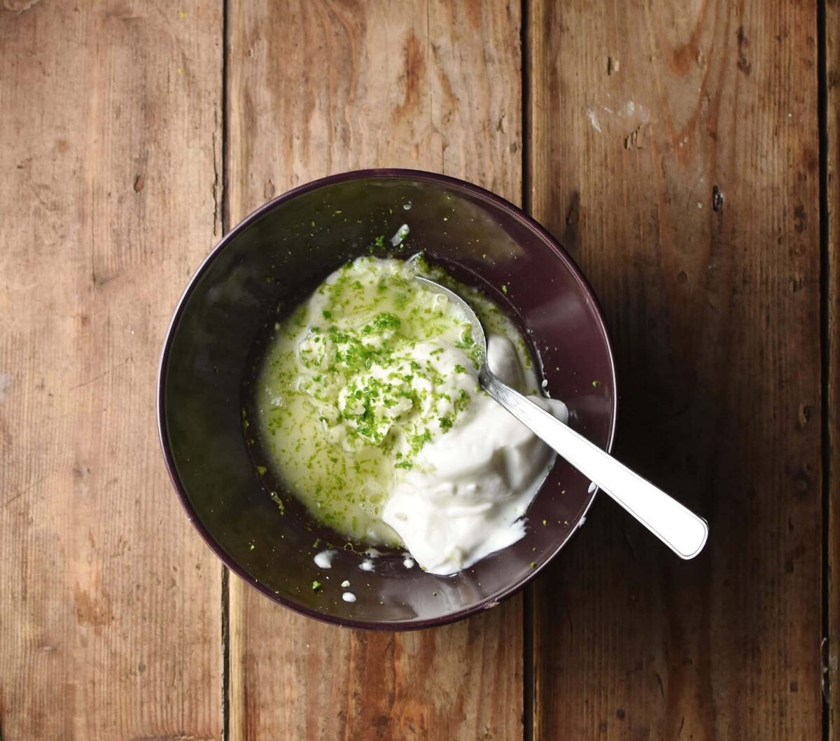 Top down view of yogurt, lime zest and oil in purple bowl with spoon.