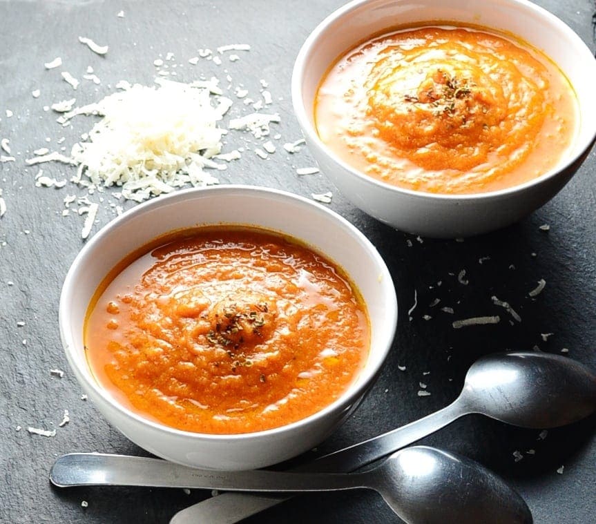 Side view of creamy tomato soup in 2 white bowls with grated cheese and 2 spoons in background.